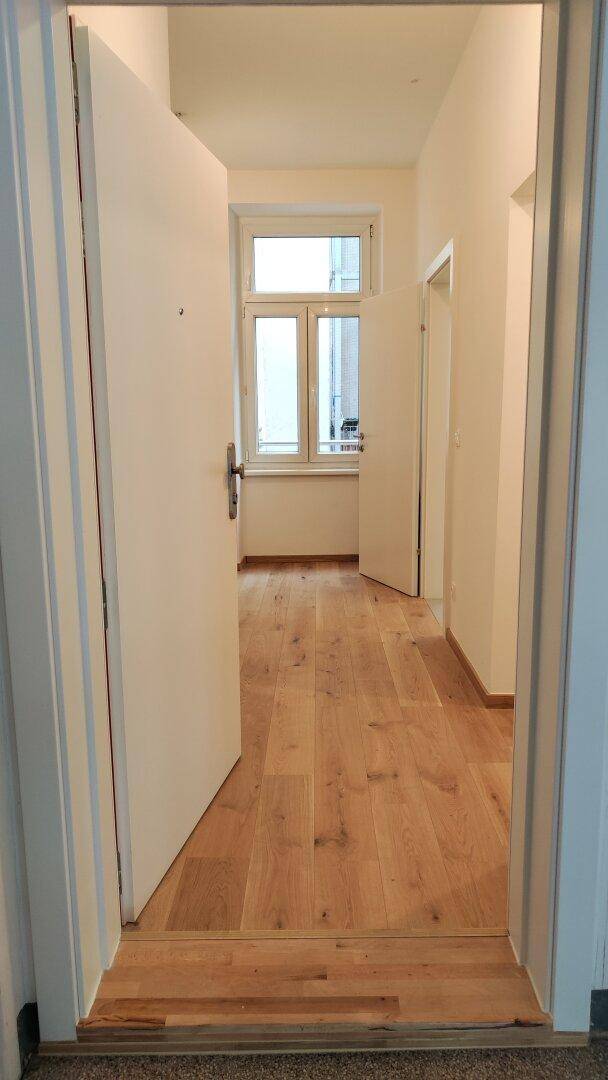 Heller Flur mit Holzboden und offener Tür, der zu weiteren Räumen mit Tageslicht führt.
