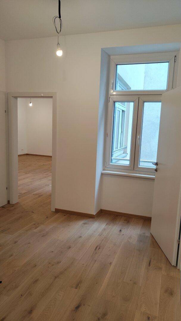 Leeres Zimmer mit hellem Holzboden und einem Fenster, das viel Tageslicht hereinlässt.