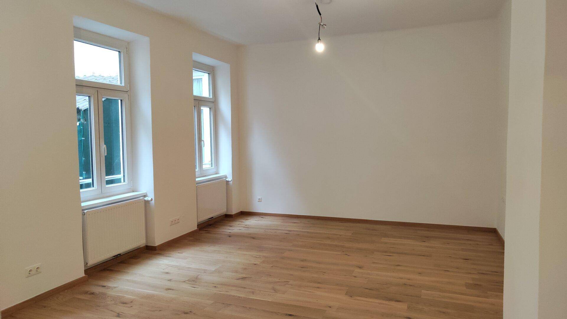 Helles, leeres Zimmer mit neuem Holzboden und zwei Fenstern, ideal für Wohn- oder Schlafbereich.