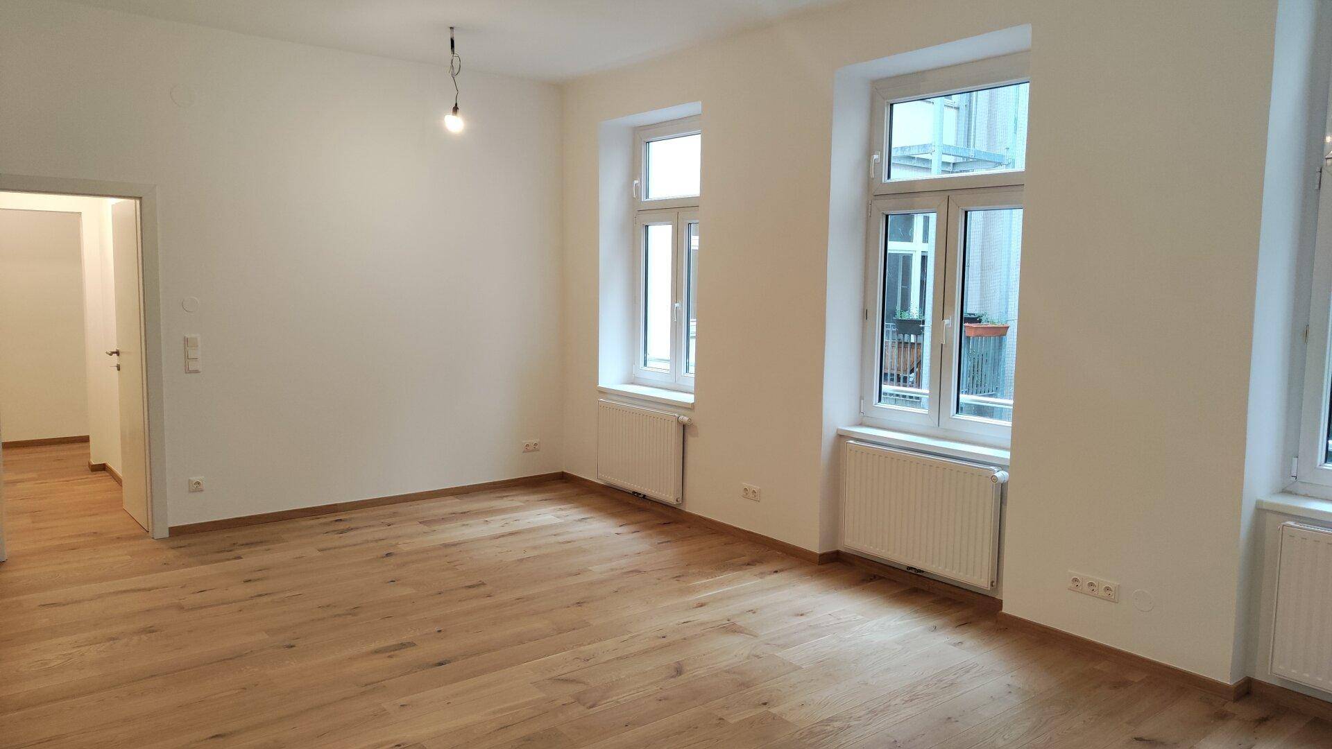 Geräumiges Zimmer mit hellem Holzboden, weißen Wänden und Fenstern, die viel Licht spenden.