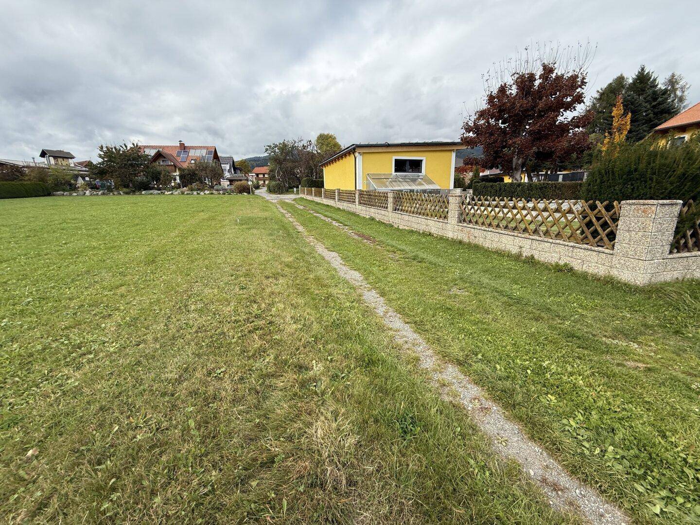 Grünes Grundstück mit Schotterweg und angrenzendem Zaun, im Hintergrund weitere Wohnhäuser sichtbar.