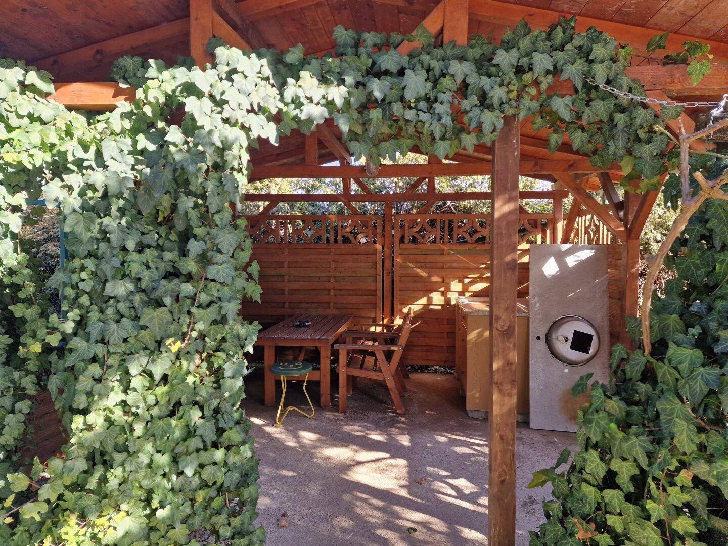 Hölzerne Pergola mit Efeu bewachsen, bietet einen schattigen Sitzbereich im Freien auf einer gepflasterten Fläche.