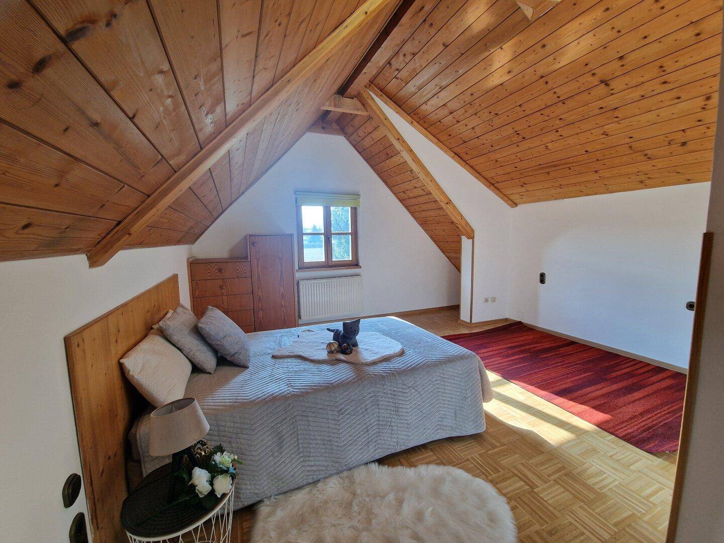 Gemütliches Schlafzimmer im Dachgeschoss mit Holzbalkendecke, Parkettboden und Fenster.