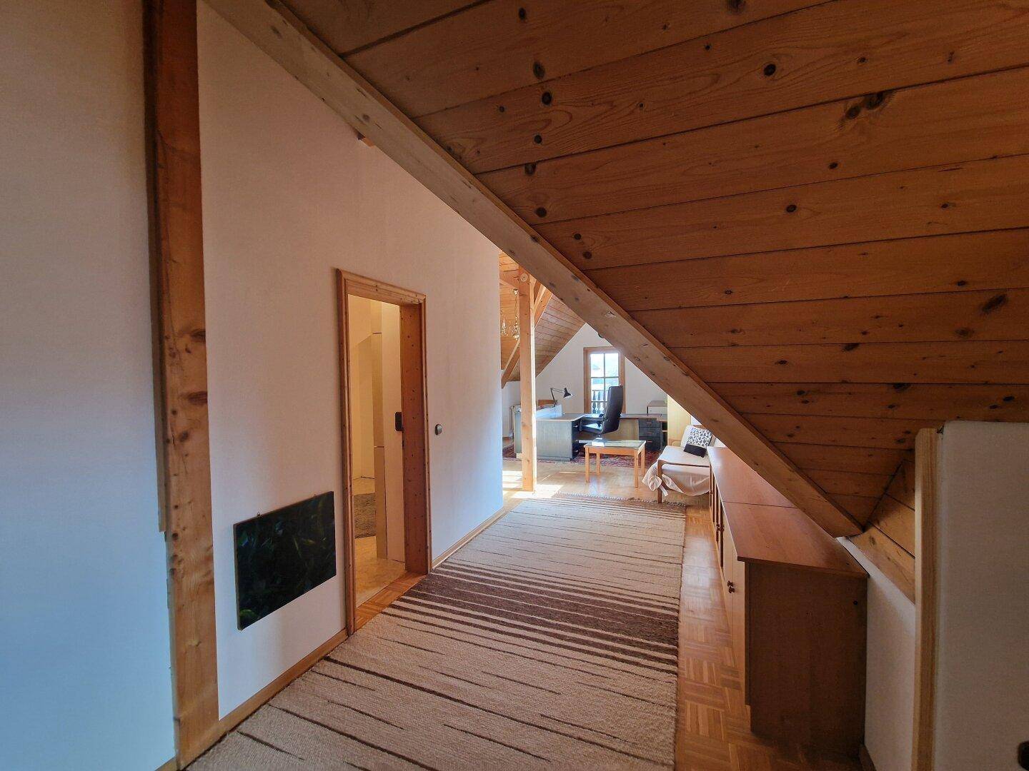 Geräumiger Flur im Dachgeschoss mit Teppichläufer, Holzbalken und Blick auf den Bürobereich.