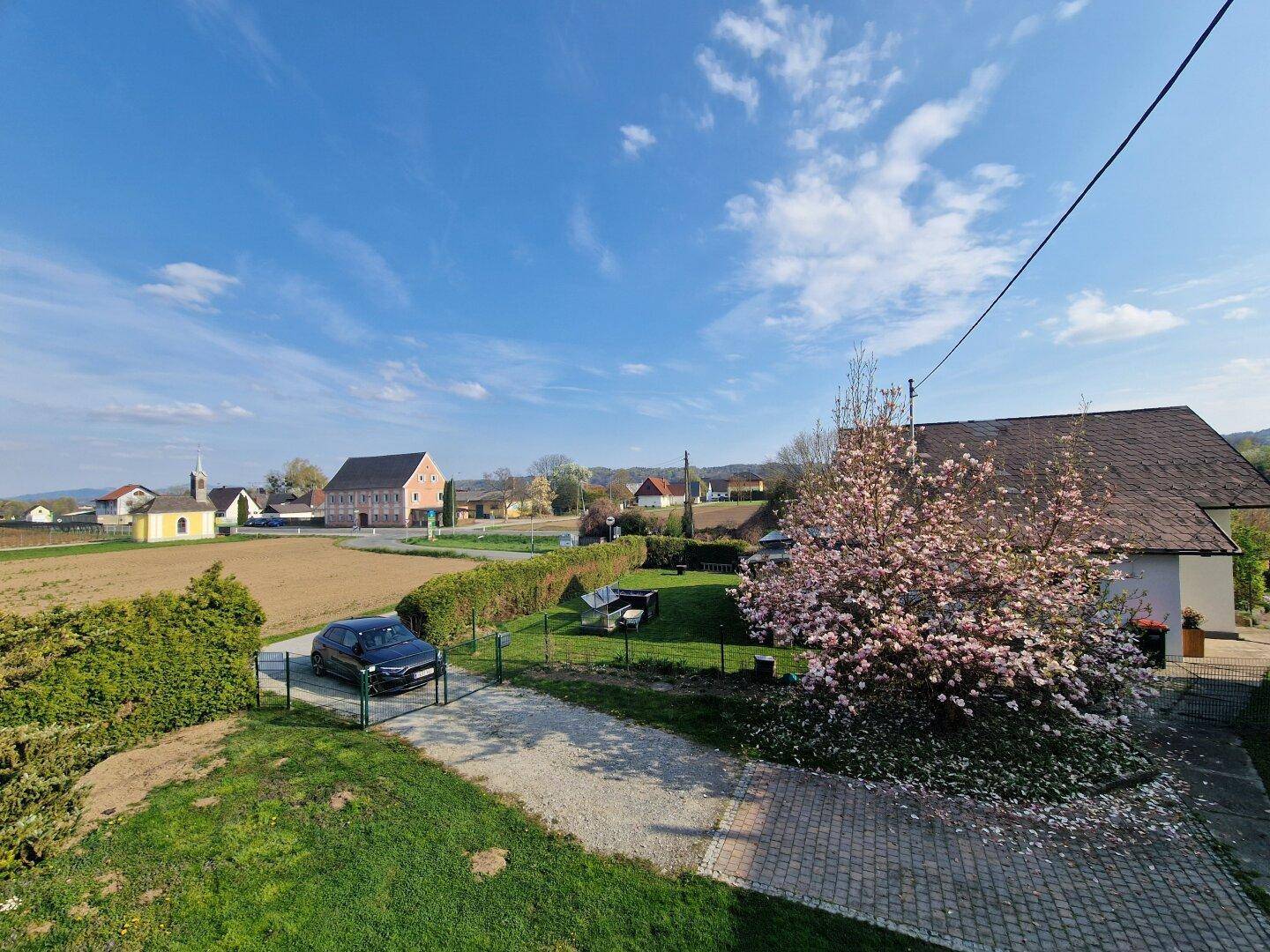 Weitläufige ländliche Umgebung mit Feldern, Häusern und blühenden Bäumen unter blauem Himmel.