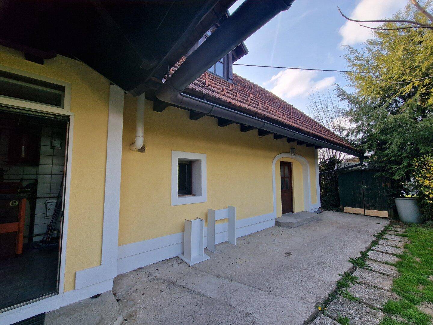 Seitenansicht des Hauses mit gelber Fassade, rotem Ziegeldach und einem Eingangsbereich mit gepflastertem Weg.