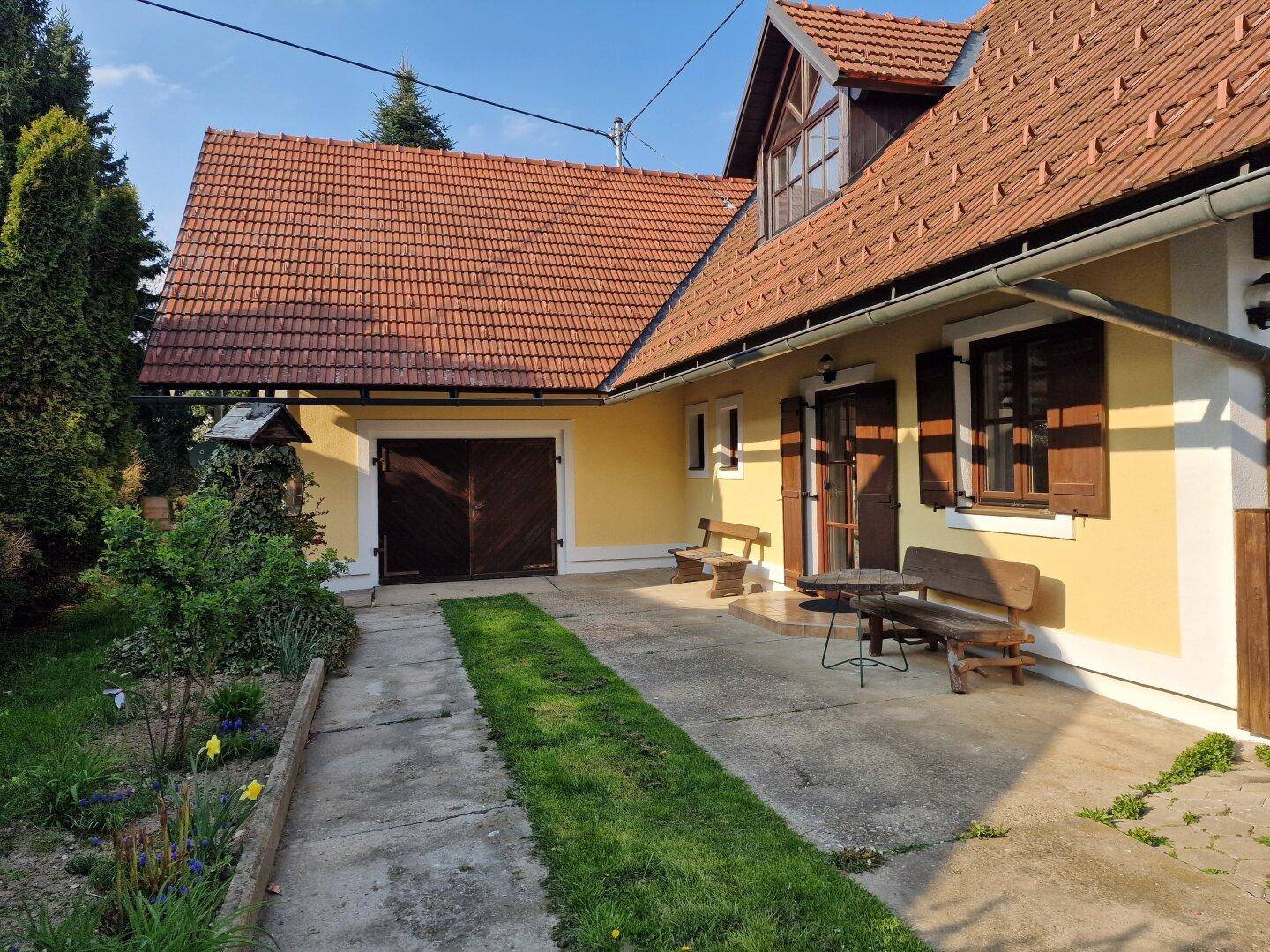 Hausfassade mit gelbem Anstrich, rotem Ziegeldach, Garage und gepflasterter Zufahrt im Grünen.