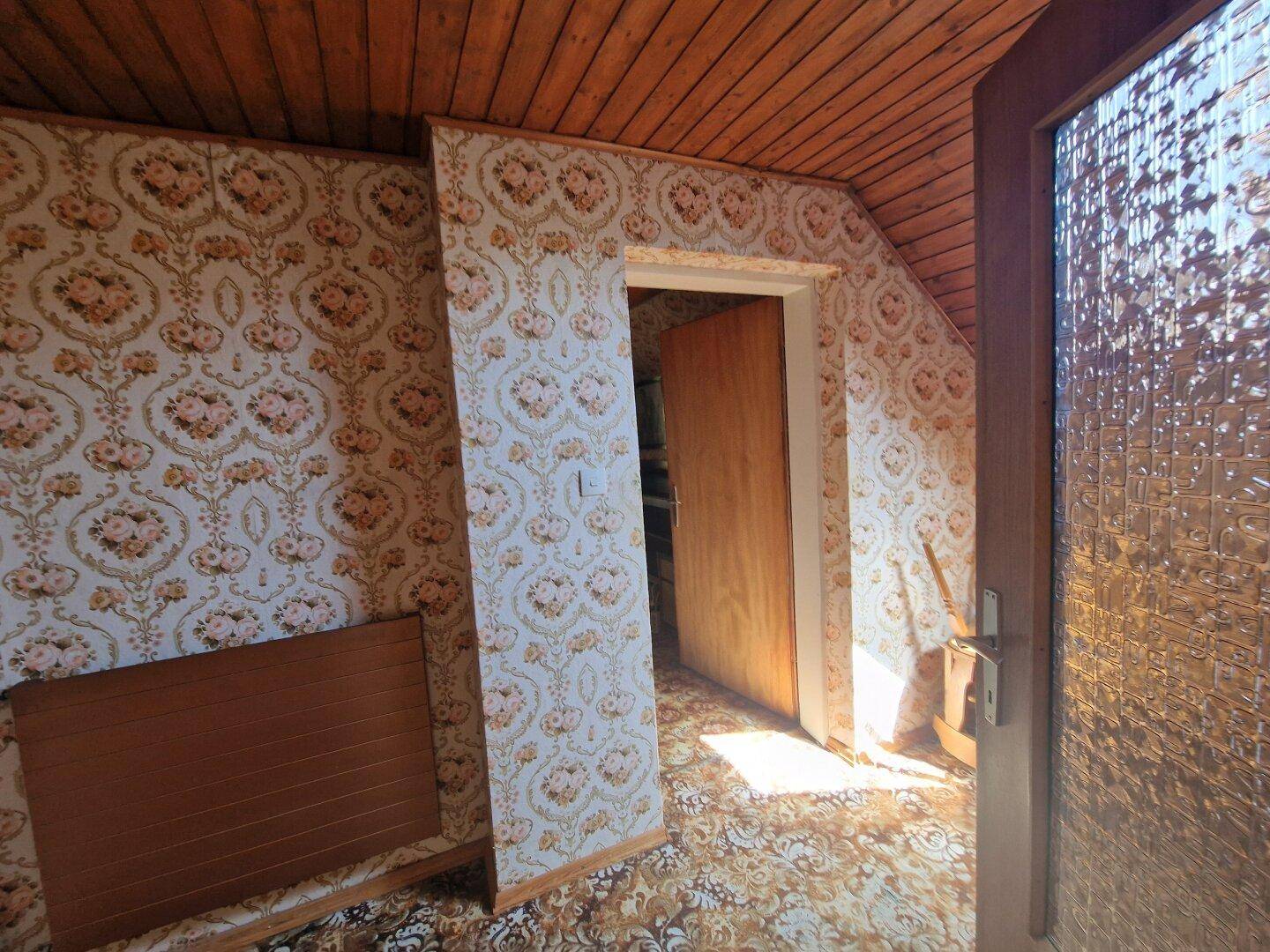 Dachgeschossraum mit gemusterter Tapete, Holztäfelung an der Decke und Tür.