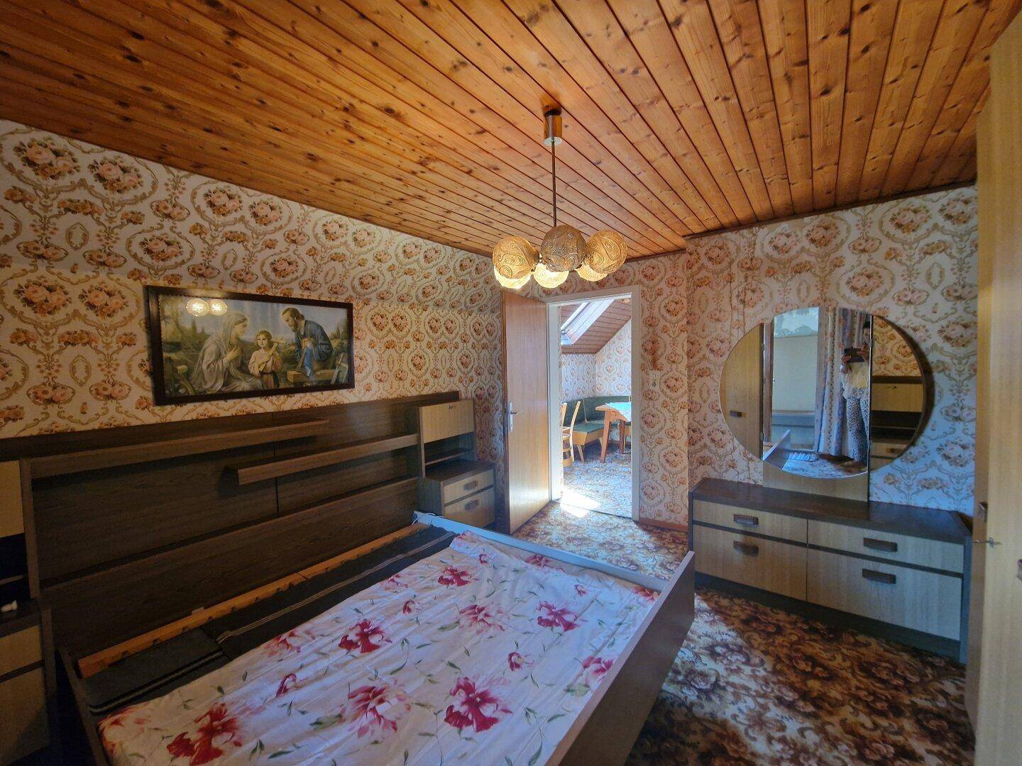 Traditionelles Schlafzimmer im Dachgeschoss mit gemusterter Tapete, Holzbett und Spiegel.
