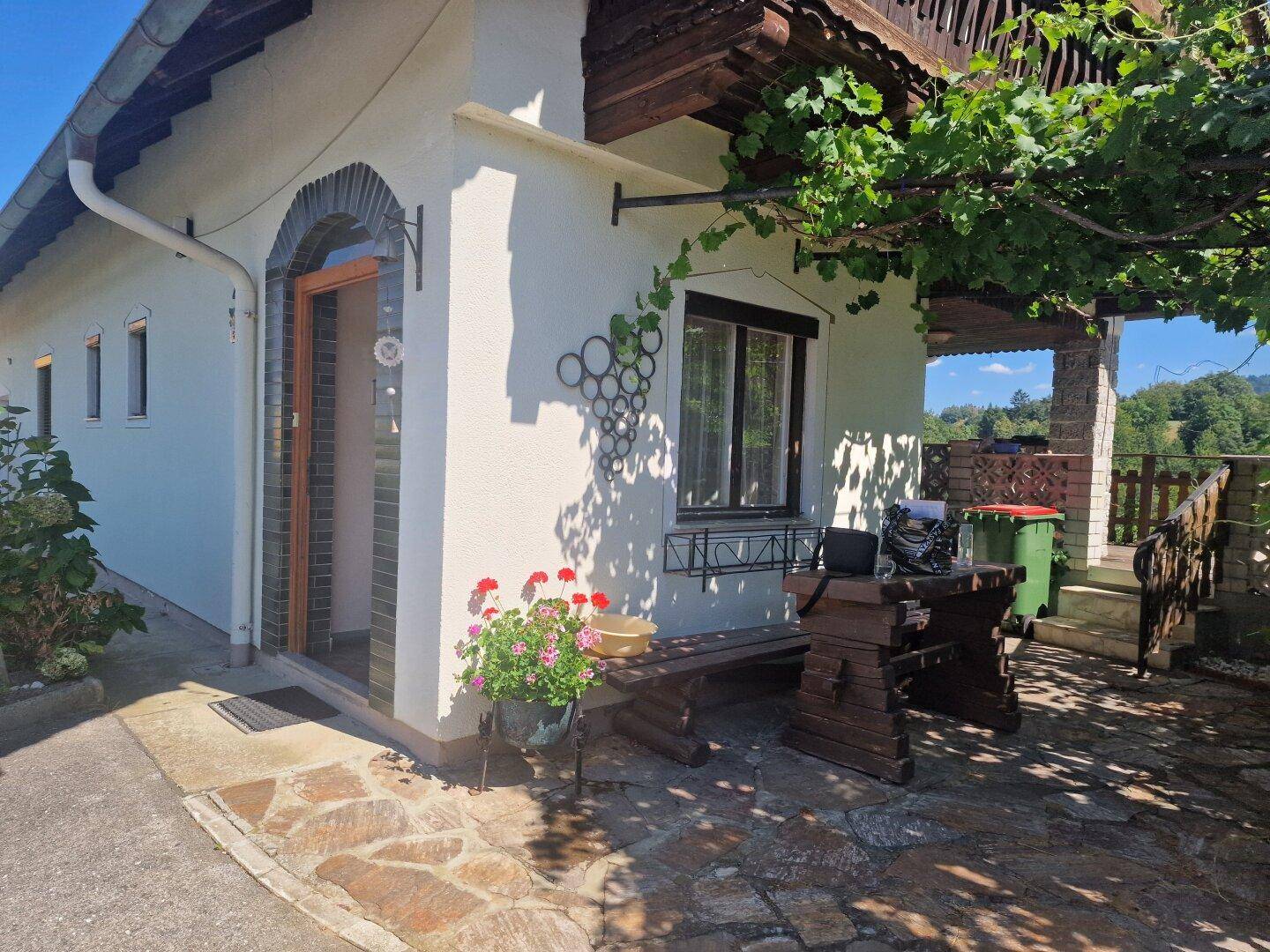 Eingangsbereich des Hauses mit einer kleinen gepflasterten Terrasse und rustikalen Holzmöbeln.