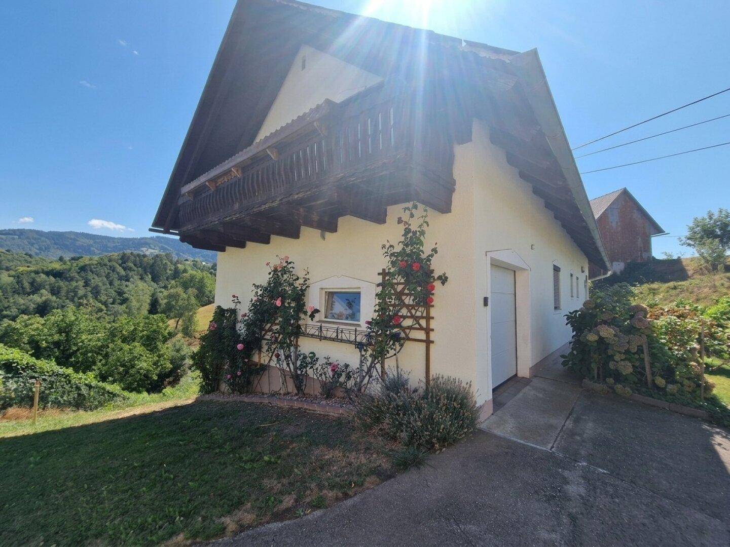 Charmantes Haus mit Balkon, Garage und blühenden Rosensträuchern vor grüner Hügellandschaft.