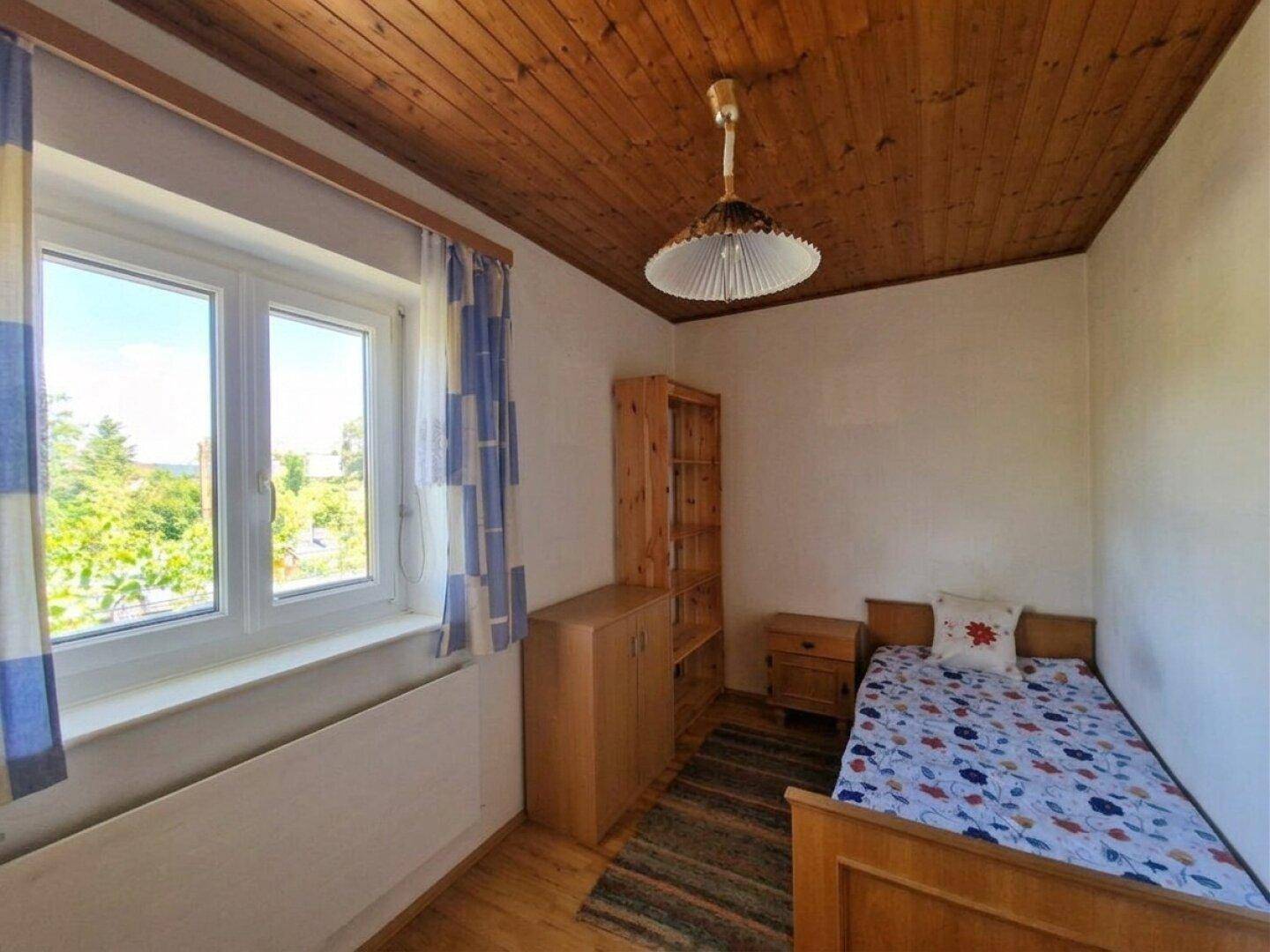 Einfaches Schlafzimmer mit Holzboden, Einzelbett und Fenster mit Blick ins Grüne.