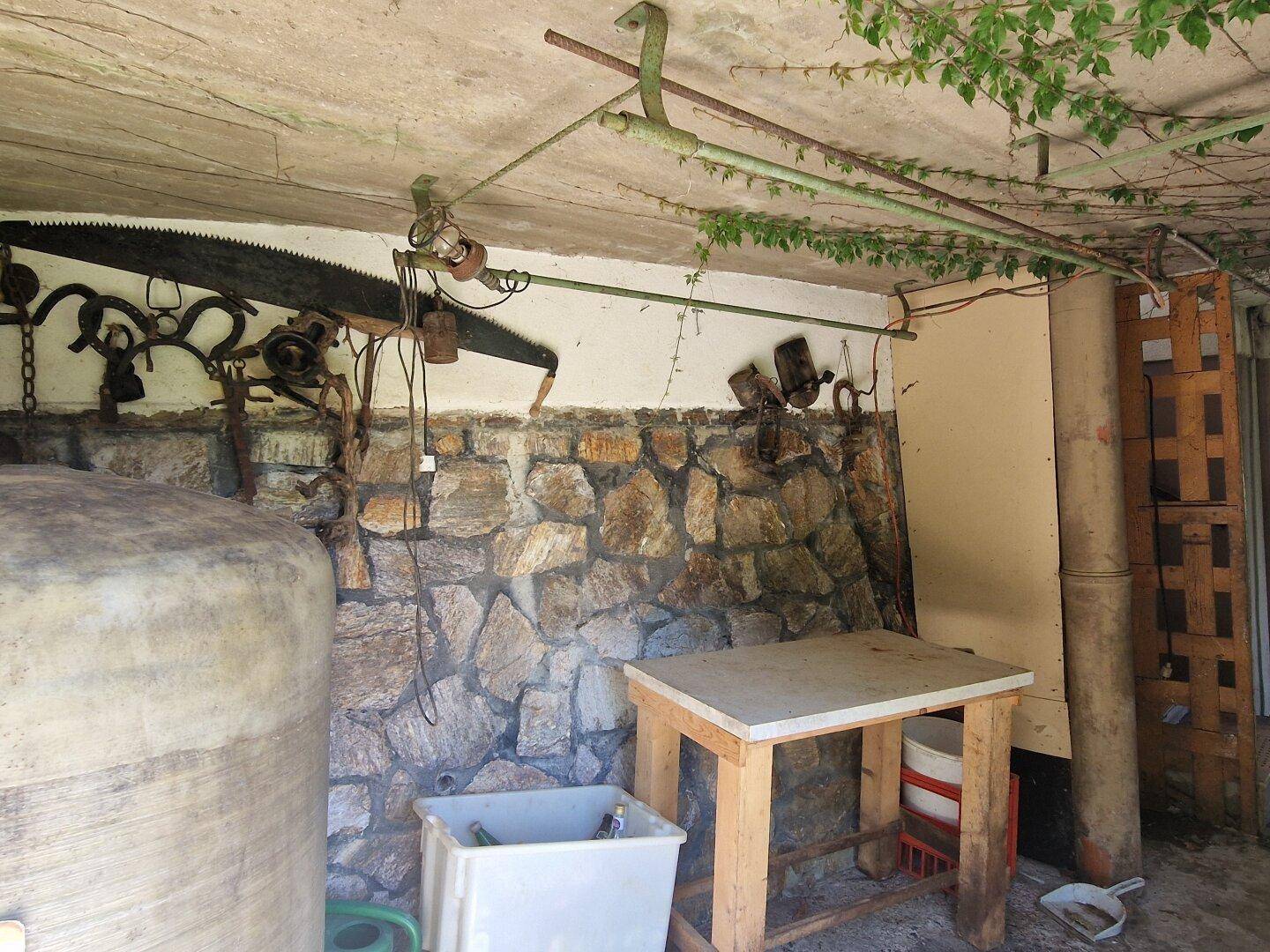 Überdachter Außenlagerbereich mit Steinmauer, alten Werkzeugen und einem großen Wassertank.