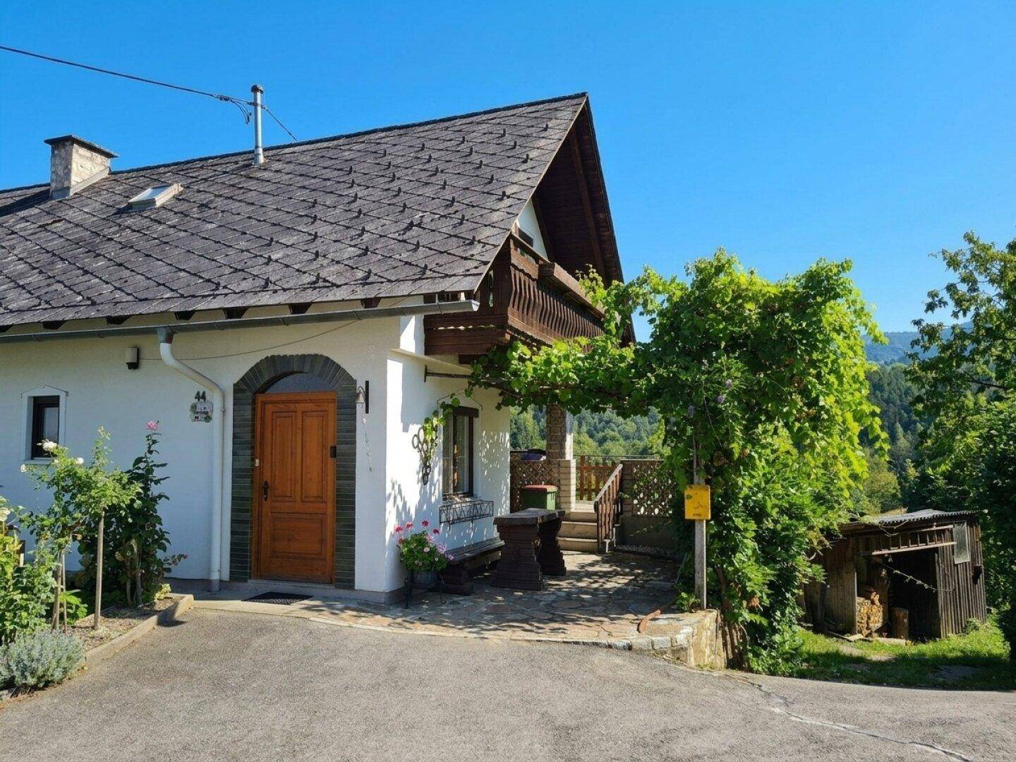 Vorderansicht des Hauses mit Eingang, kleiner Terrasse und einem Holzschuppen im Hintergrund.