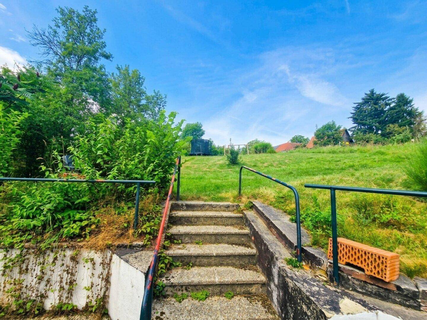 Steintreppe führt einen grünen, bewachsenen Hang hinauf zu einem oberen Gartenbereich.