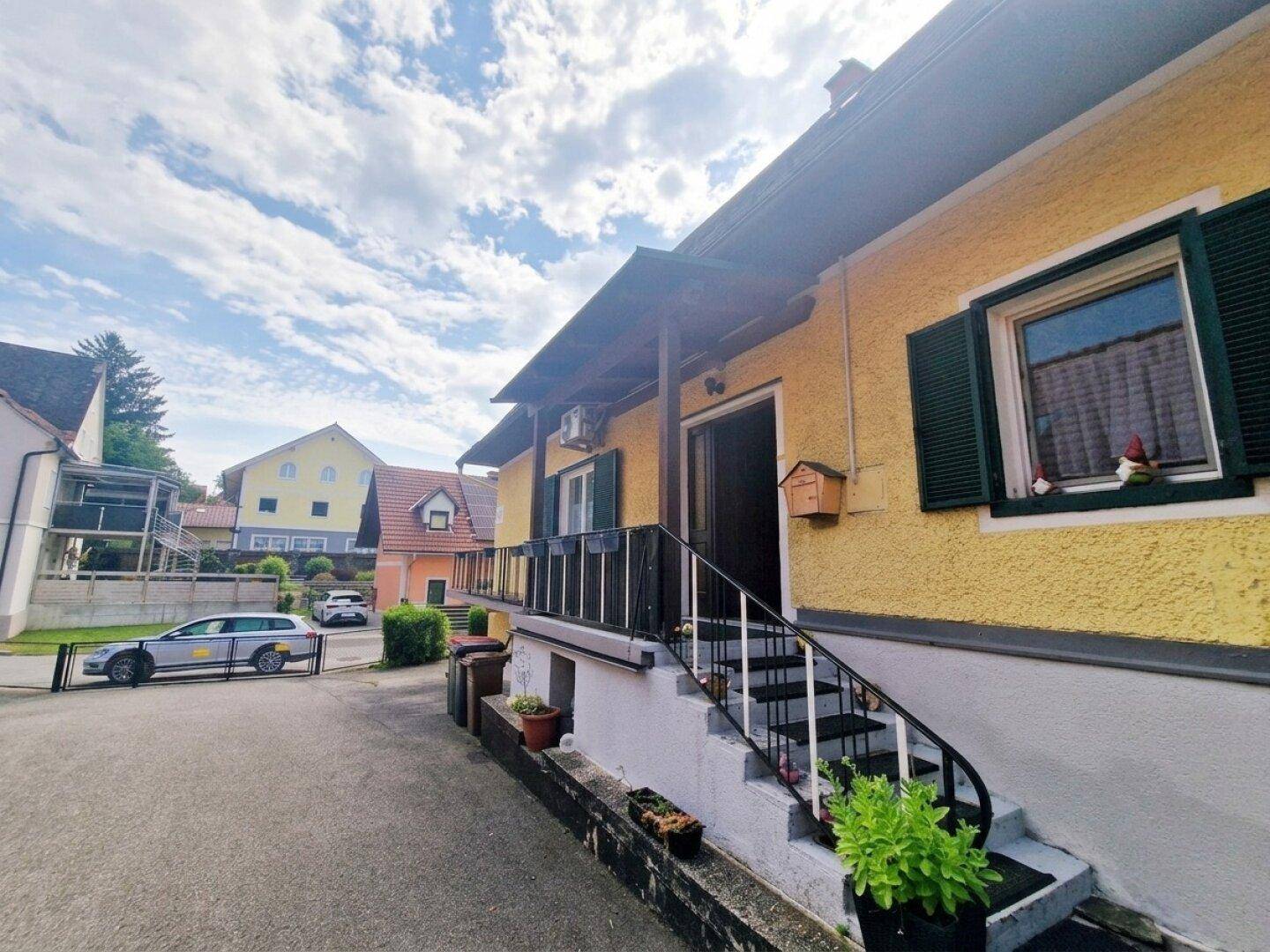 Eingangsbereich des gelben Hauses mit überdachtem Podest und grünen Fensterläden.