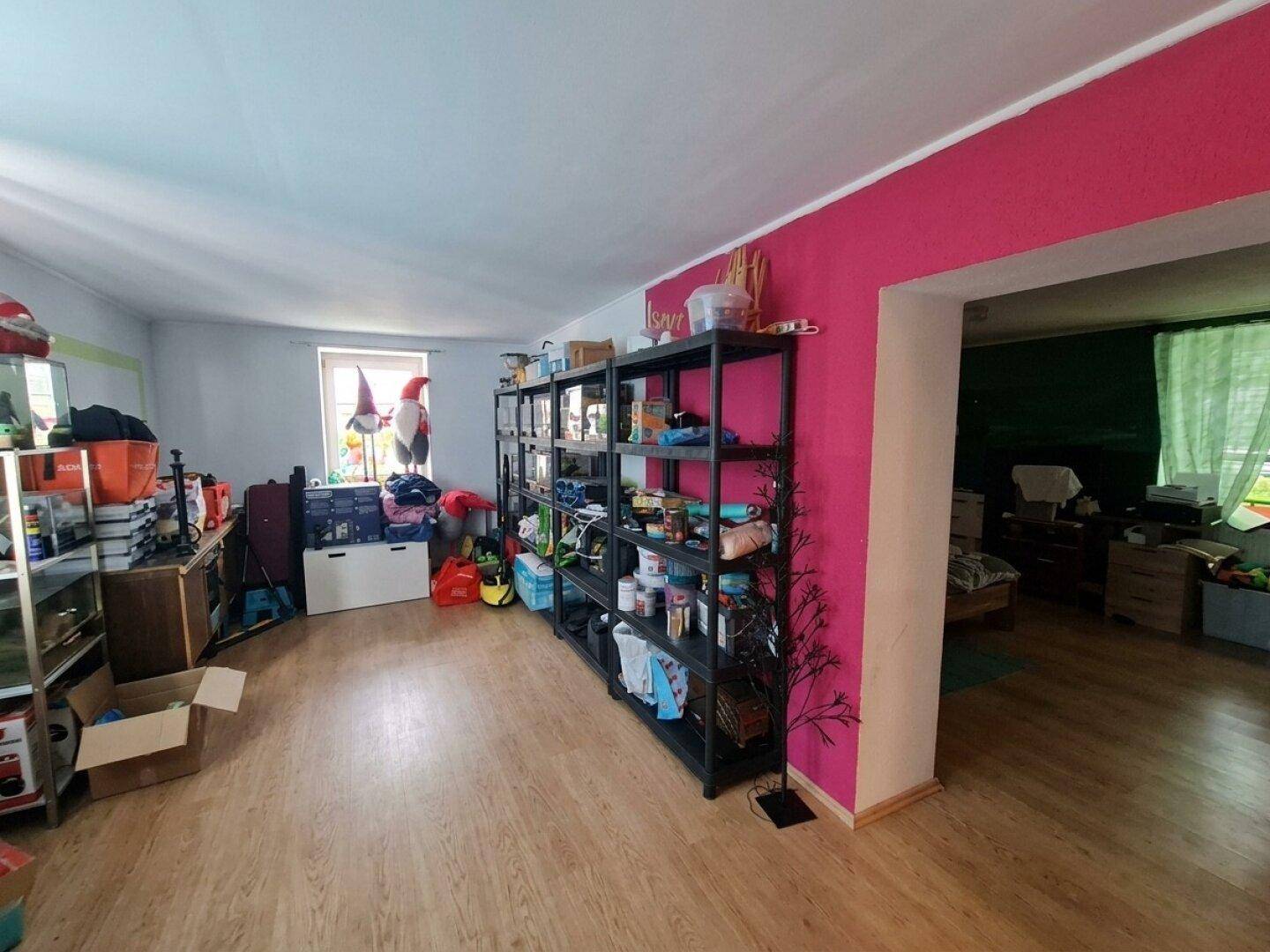 Geräumiger Raum mit pinkfarbener Wand, Regalen und Zugang zu einem weiteren Zimmer.