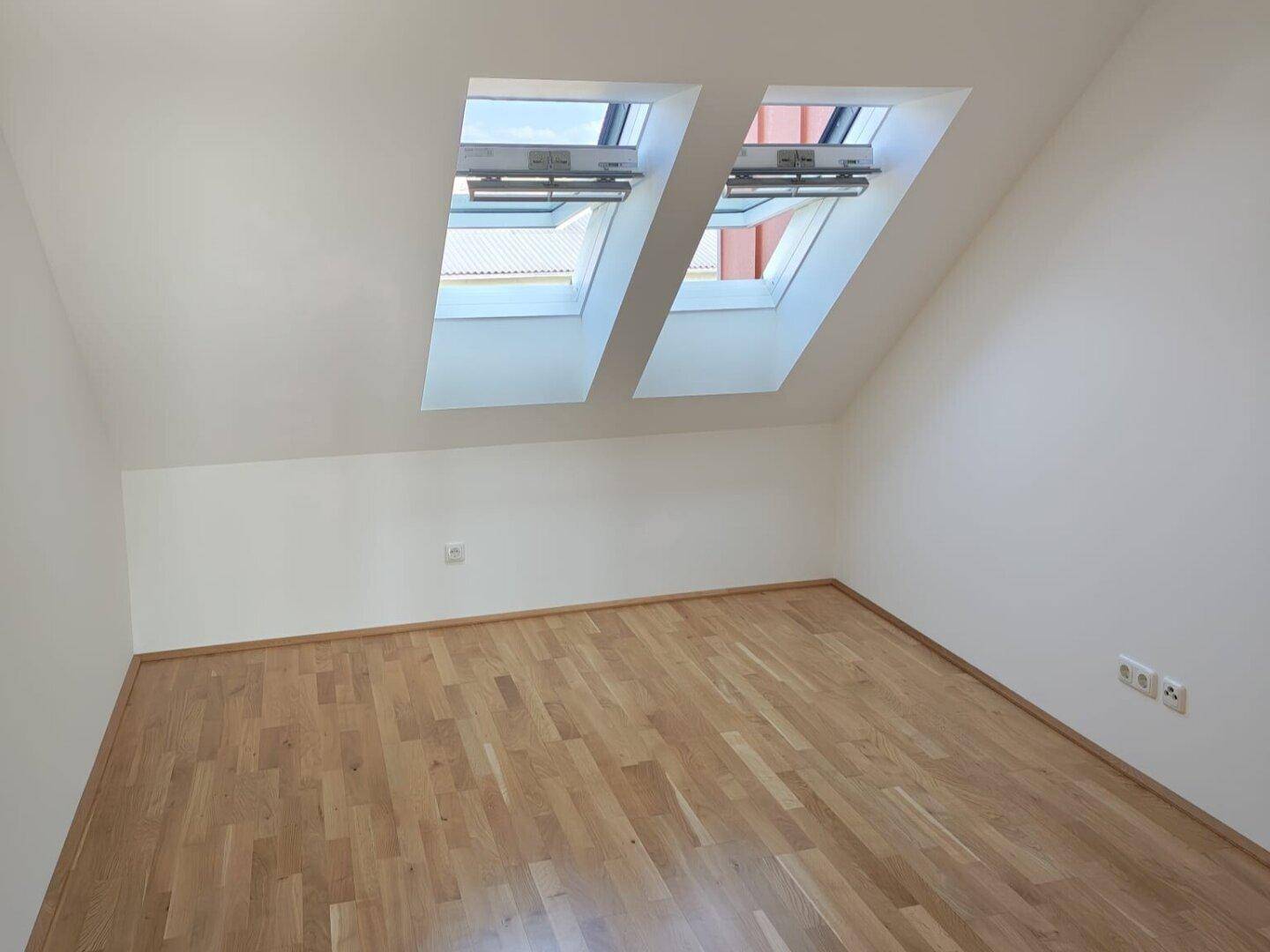 Helles Zimmer im Dachgeschoss mit zwei Dachfenstern und hellem Holzparkettboden.