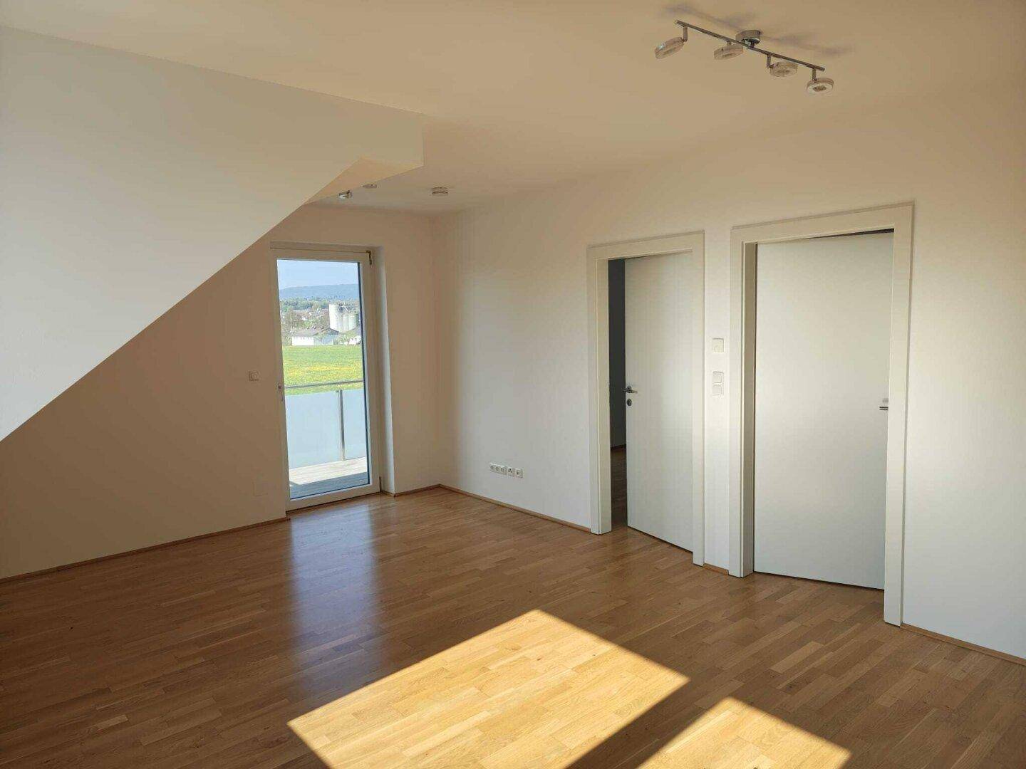 Heller Wohnbereich mit Holzboden, Zugang zu einem Balkon und Blick ins Grüne.