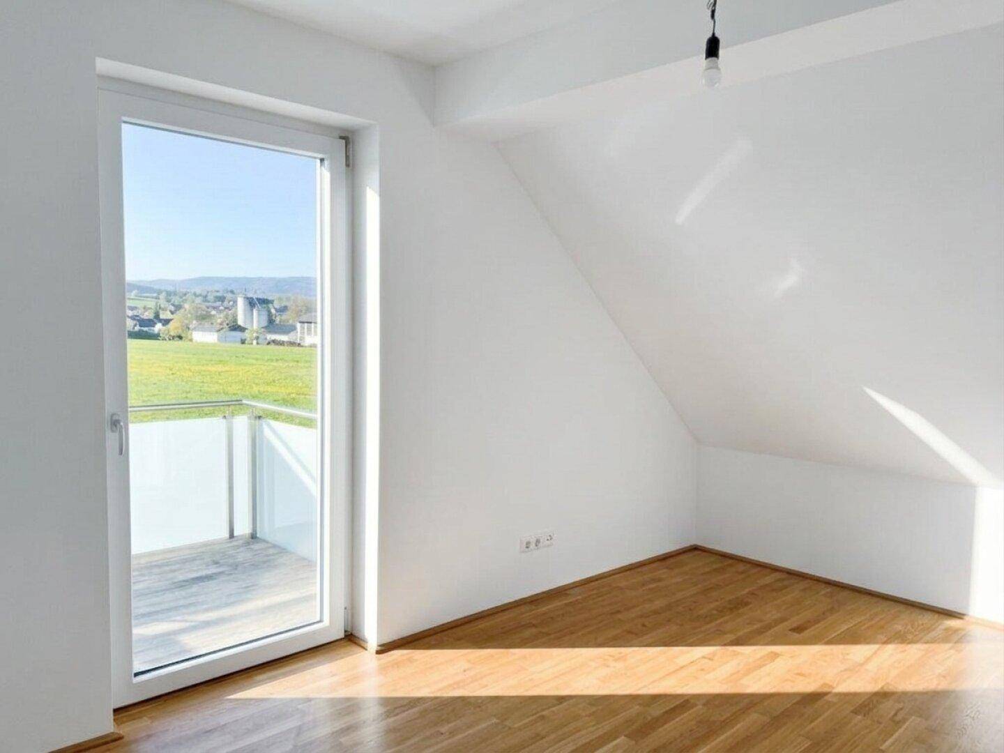 Lichtdurchflutetes Zimmer mit Holzboden und großem Fenster mit Zugang zum Balkon.