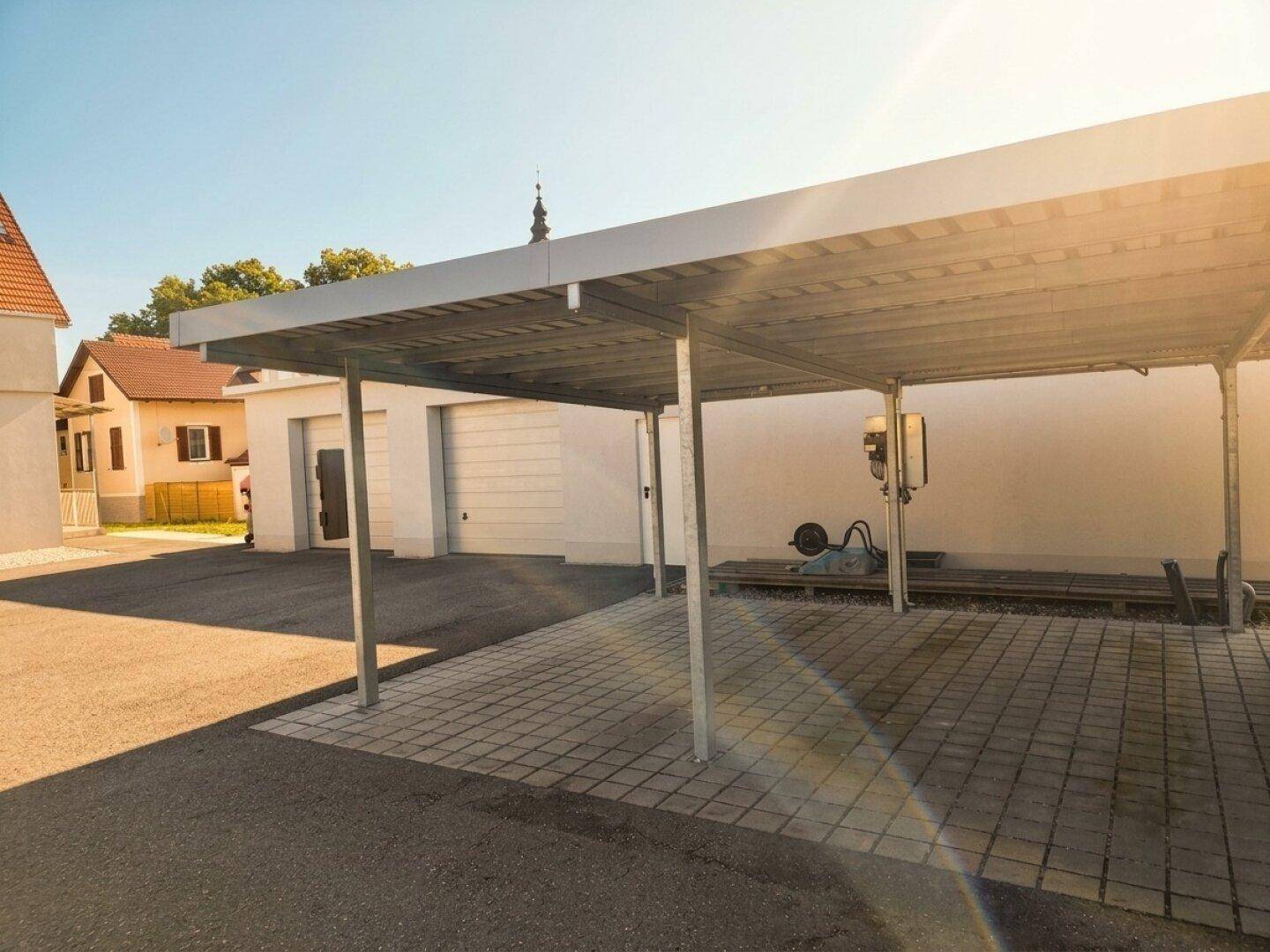 Überdachter Parkplatz (Carport) mit Ladestation für Elektrofahrzeuge und Garagen im Hintergrund.