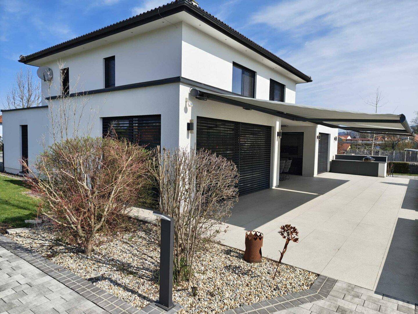 Moderne Hausfassade mit großer Auffahrt, Markise und gepflegter Vorgartenbepflanzung.
