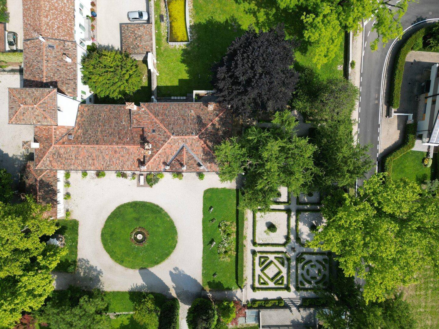 Draufsicht auf die historische Villa mit ihrem markanten Dach, der kreisförmigen Auffahrt und dem geometrischen Garten.