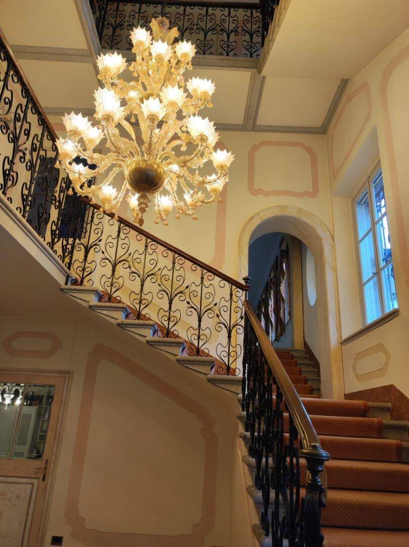 Die elegante Treppe mit Teppichläufer und einem großen Kronleuchter, umgeben von historischen Wandmalereien.
