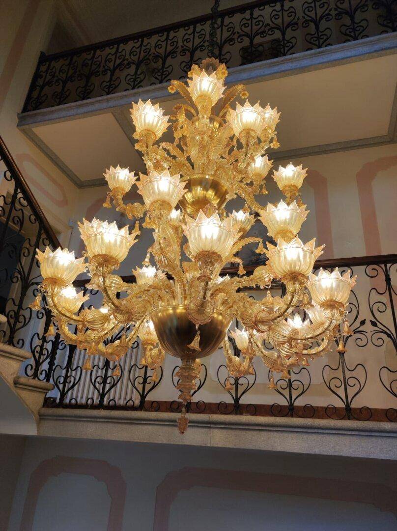 Ein prunkvoller goldener Kronleuchter hängt majestätisch im hohen Treppenhaus mit schmiedeeisernem Geländer.
