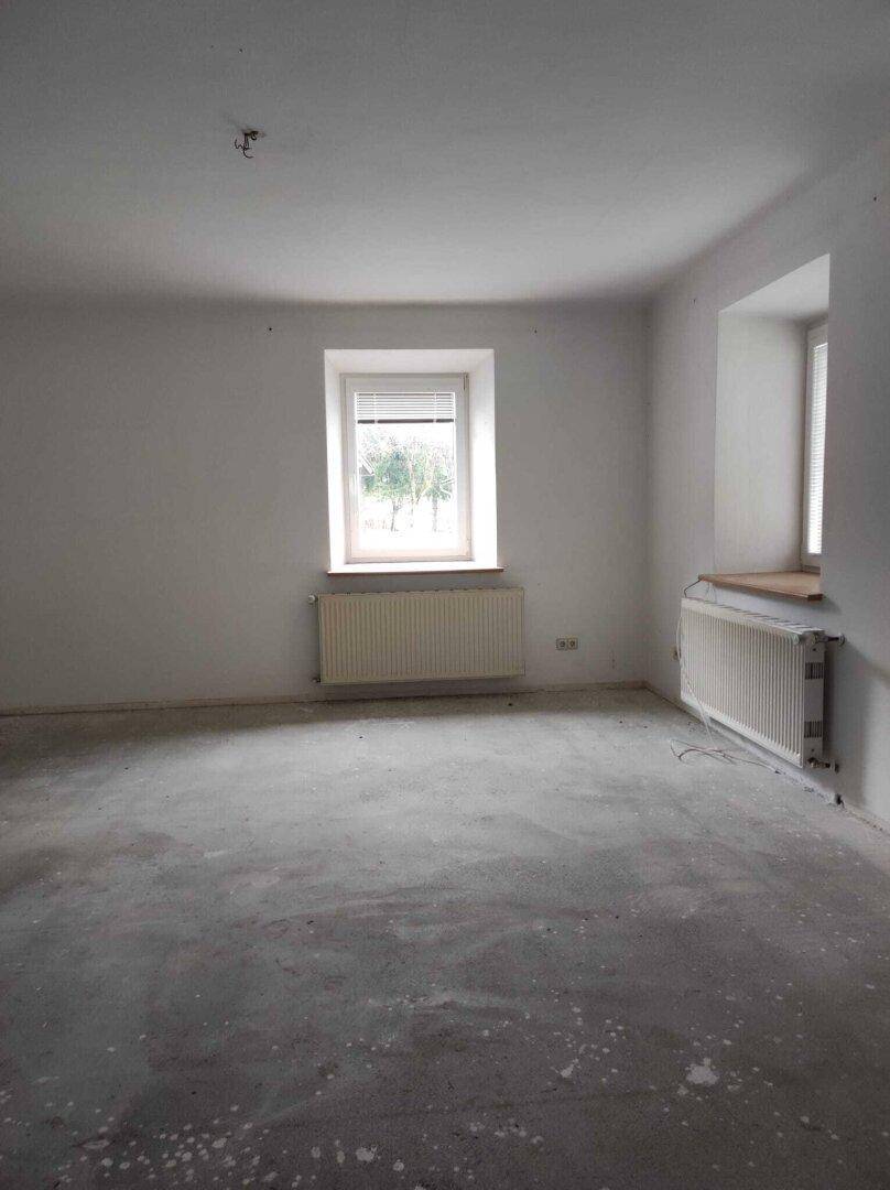 Heller, leerer Raum mit zwei Fenstern, Heizkörpern und Betonboden, wartet auf neue Bewohner.