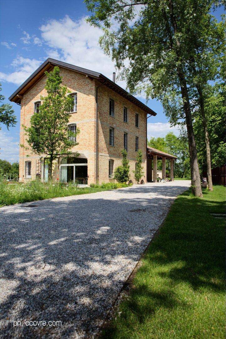 Seitenansicht des Ziegelsteinhauses mit langem Kiesweg, umgeben von hohen Bäumen und viel Grün.