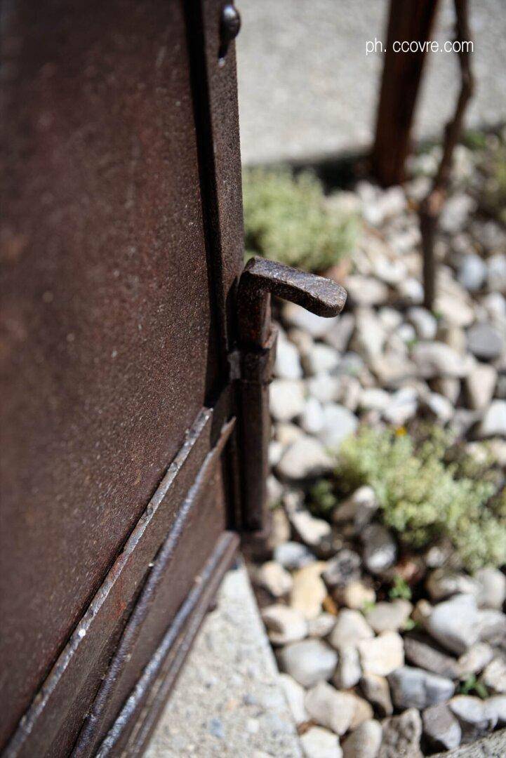 Rostiger Metallriegel eines Tores, umgeben von Kies und kleinen Pflanzen im Sonnenlicht.