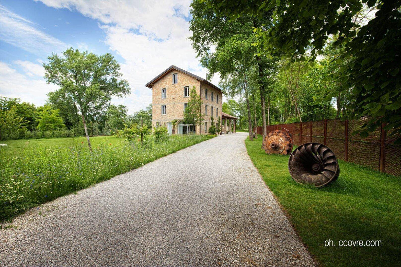 Das Ziegelsteinhaus aus der Ferne, mit langem Kiesweg und dekorativen historischen Maschinenteilen.