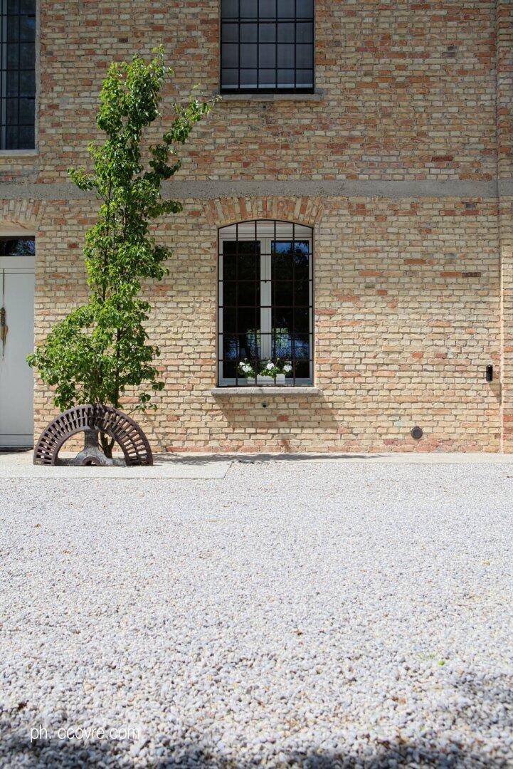 Fassade eines Backsteingebäudes mit einem kleinen Baum, vergittertem Fenster und Kiesfläche davor.