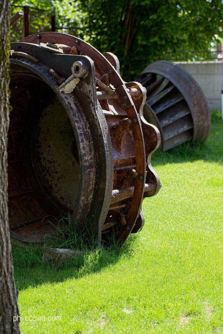 Große rostige Industriemaschinenteile als dekorative Elemente in einem grünen Gartenbereich.