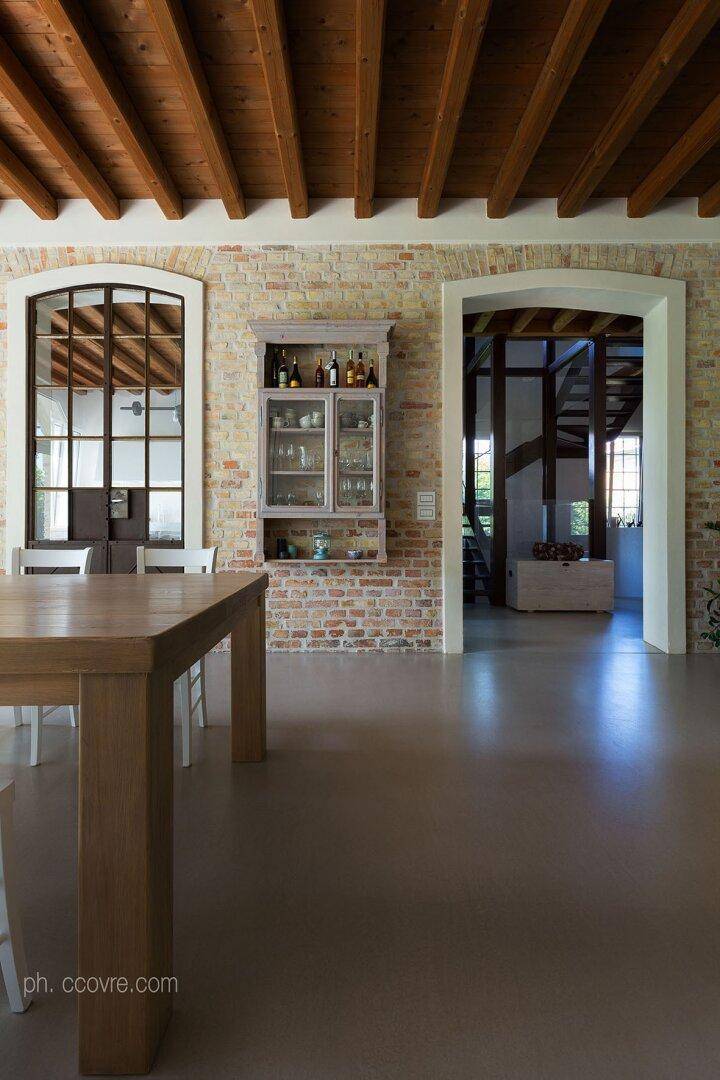 Essbereich mit Holztisch, Backsteinwand, Hängeschrank und offenem Durchgang zu einem Treppenhaus.