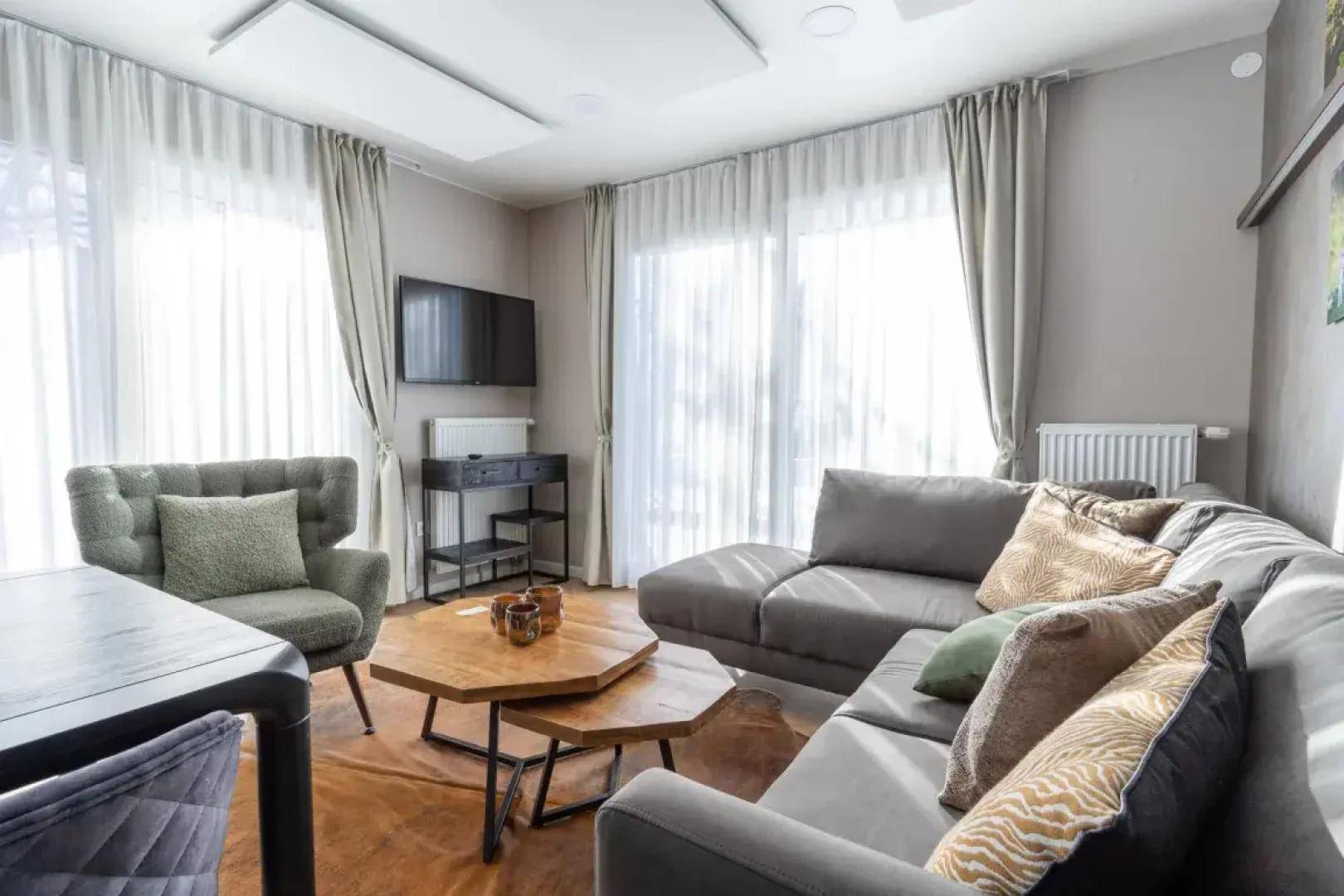 Gemütliches Wohnzimmer mit großem Ecksofa, Holztischen und viel Tageslicht durch die breiten Fenster.