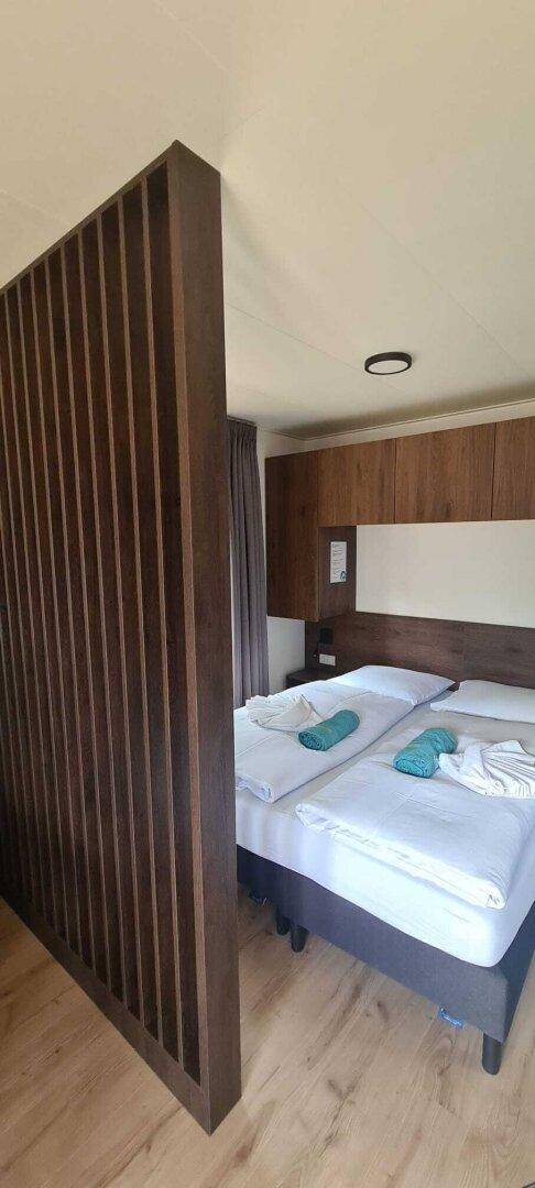 Schlafbereich mit Doppelbett und Holzwandverkleidung, abgetrennt durch eine Lamellenwand.