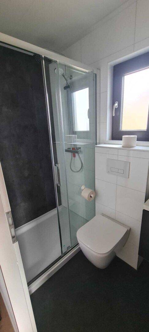 Modernes Badezimmer mit Duschkabine, wandhängendem WC und Fenster für Tageslicht.