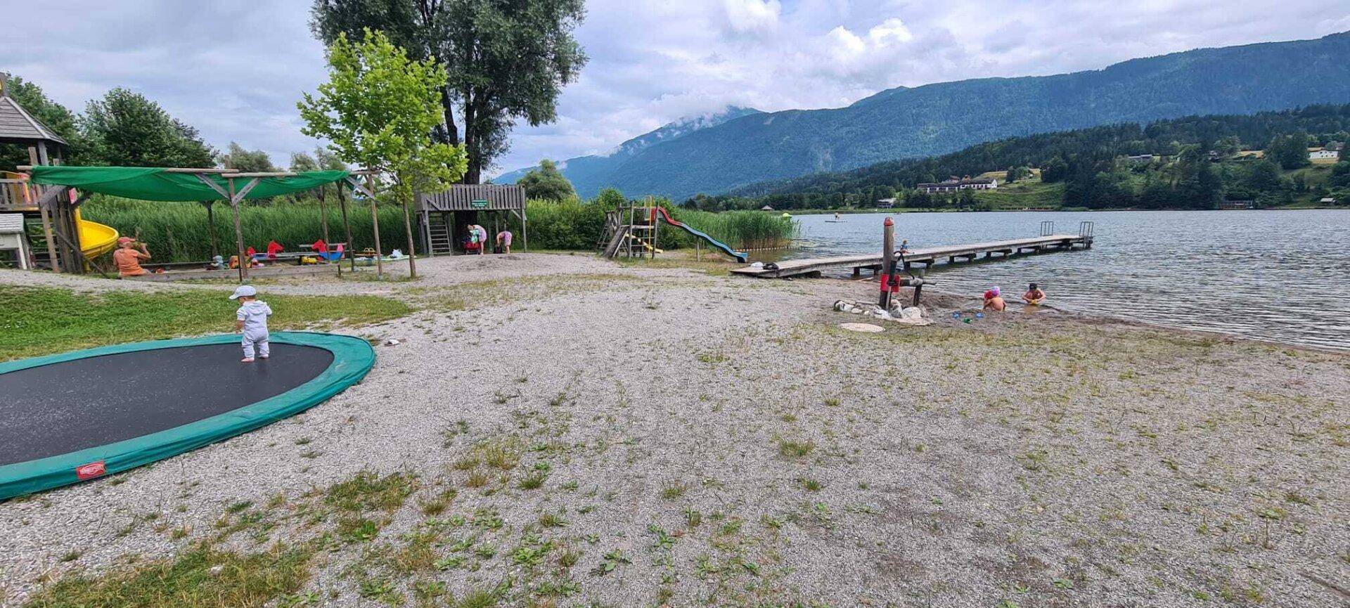 Spielbereich am Seeufer mit Trampolin, Rutsche und Steg, ideal für Familien.