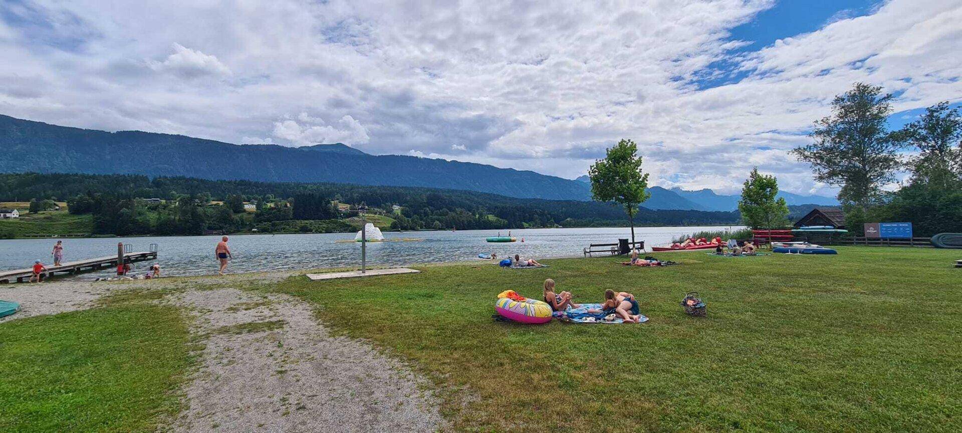 Badestrand am See mit Badegästen, Liegewiese und beeindruckendem Bergpanorama.