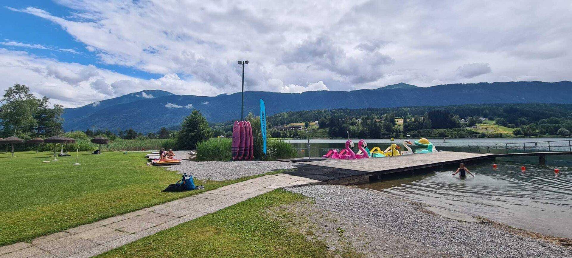 Lebhaftes Seeufer mit bunten Tretbooten, Liegewiese und Bergkulisse im Hintergrund.