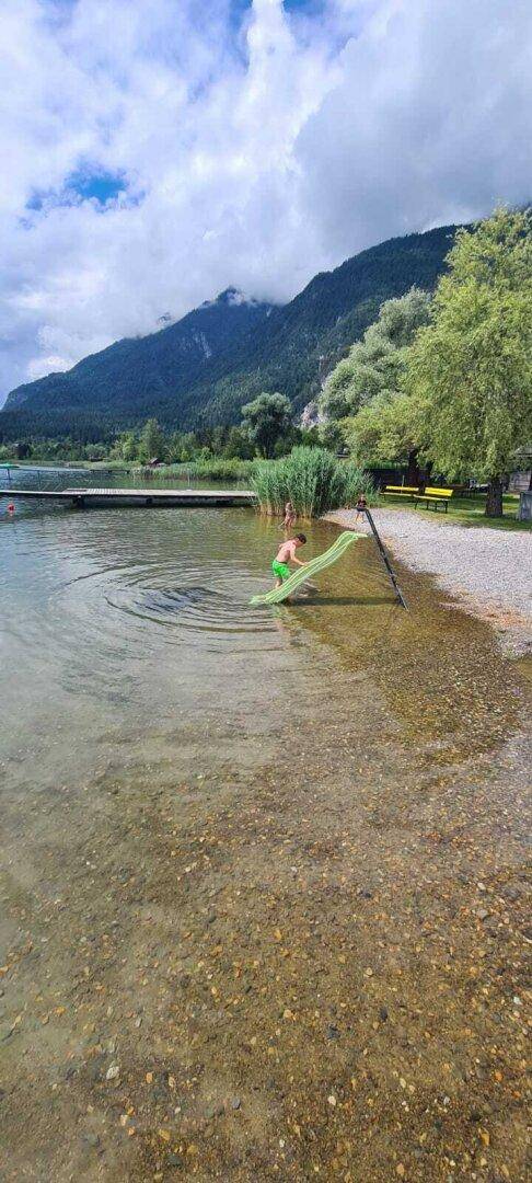 Kind spielt am flachen Seeufer mit einer kleinen Rutsche und Schilf vor Berglandschaft.