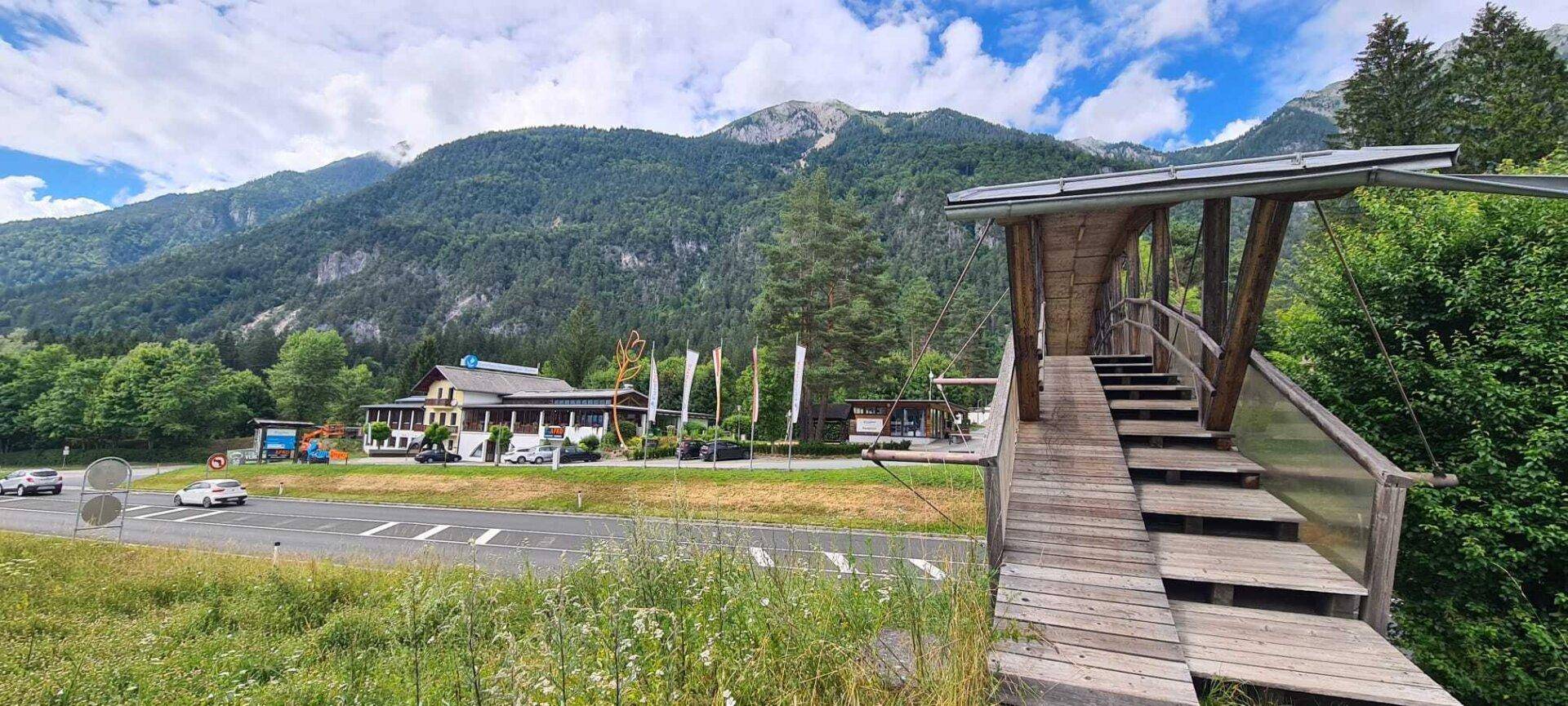 Hauptgebäude des Ferienparks mit Zufahrtsstraße und einer Holzbrücke vor Bergkulisse.