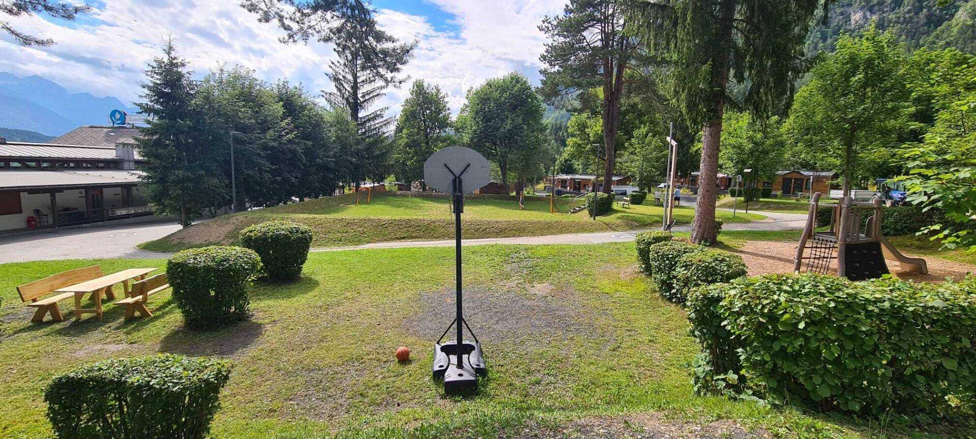Spielbereich mit Basketballkorb und Picknicktisch, umgeben von Hecken und Bäumen.