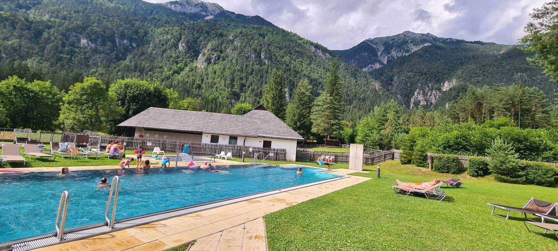 Außenpoolbereich mit Liegewiese und Badegästen, umgeben von einer malerischen Berglandschaft.