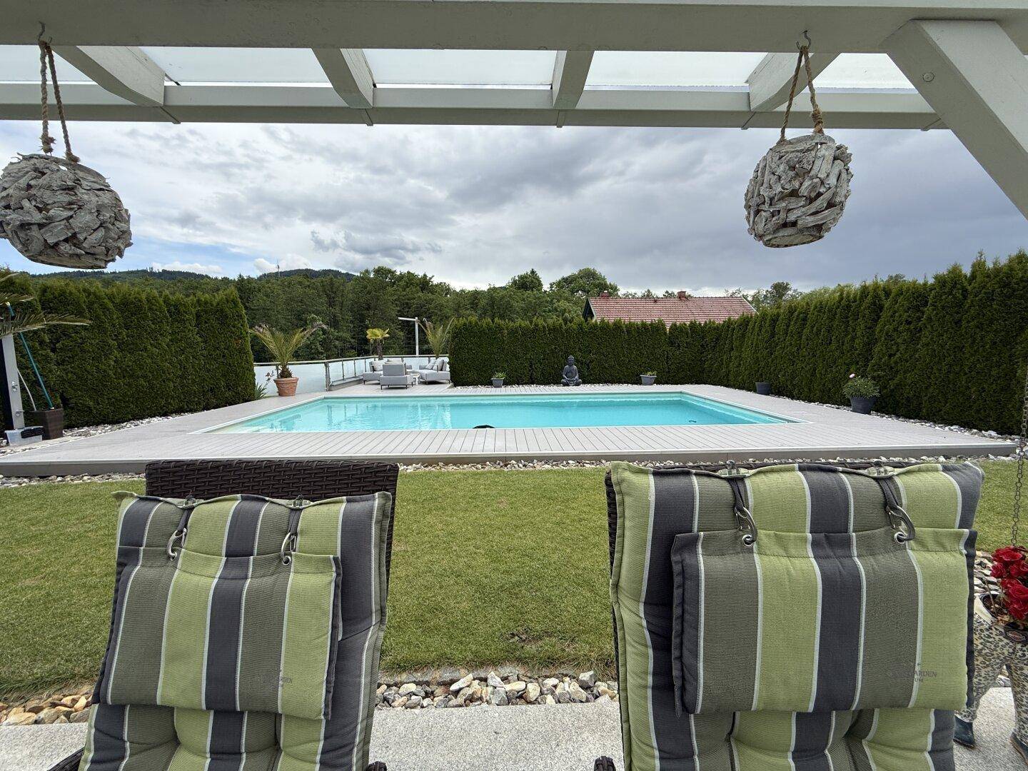 Blick von der überdachten Terrasse auf den Swimmingpool und den grünen Garten.