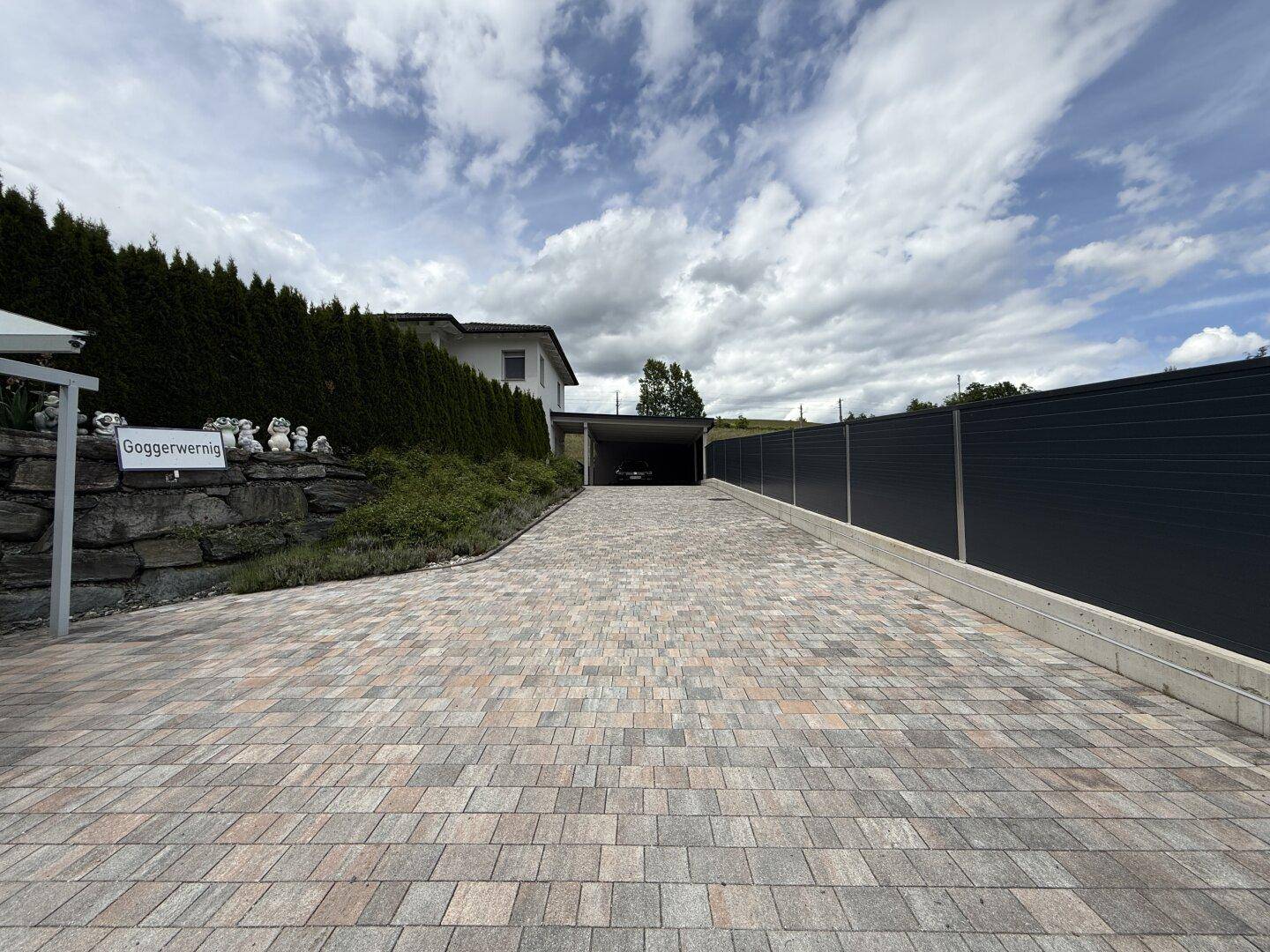 Lange, gepflasterte Auffahrt führt zum Haus mit Carport und umgebender Hecke.