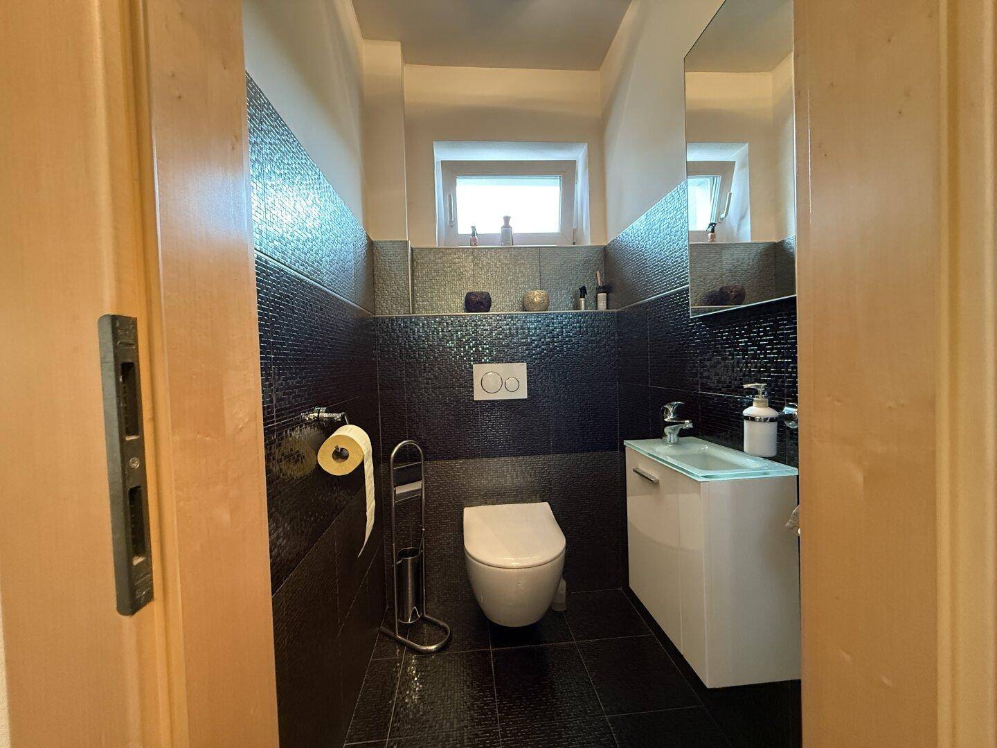 Modernes Gäste-WC mit dunklen Fliesen, wandhängendem WC und kleinem Waschbecken.