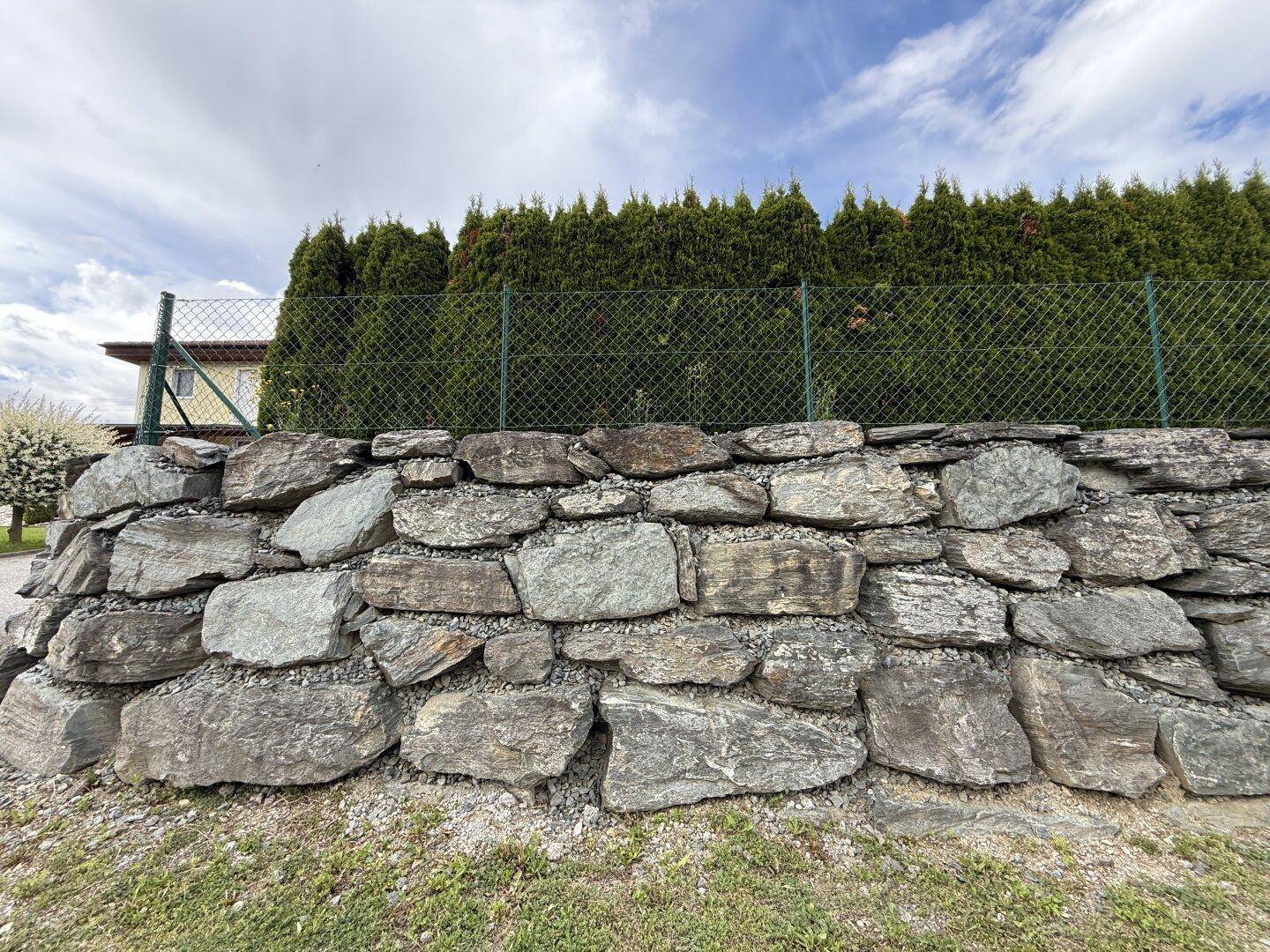 Robuste Steinmauer mit grüner Hecke im Hintergrund unter bewölktem Himmel.
